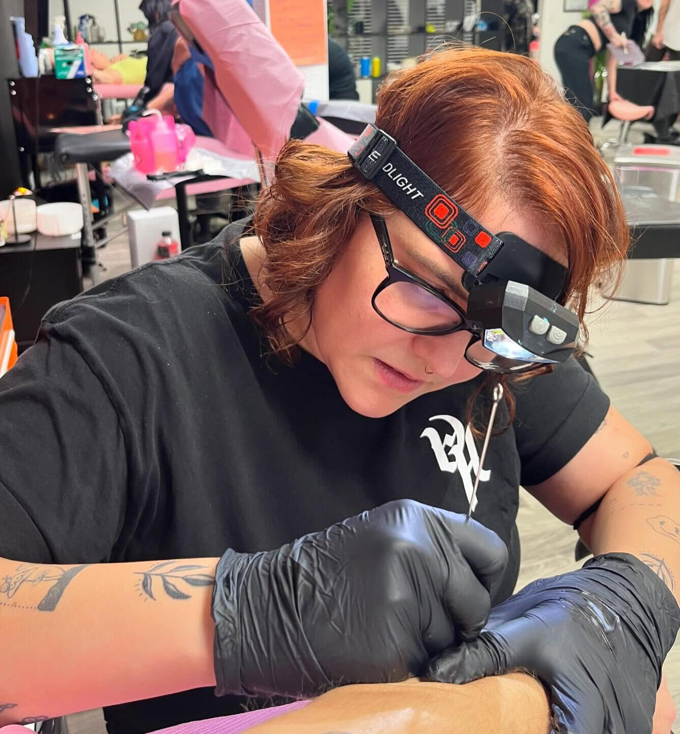 Howdy Slowpoke Tattoo Artist A person wearing glasses and a headlamp, with gloves on, tattooing someone's arm
